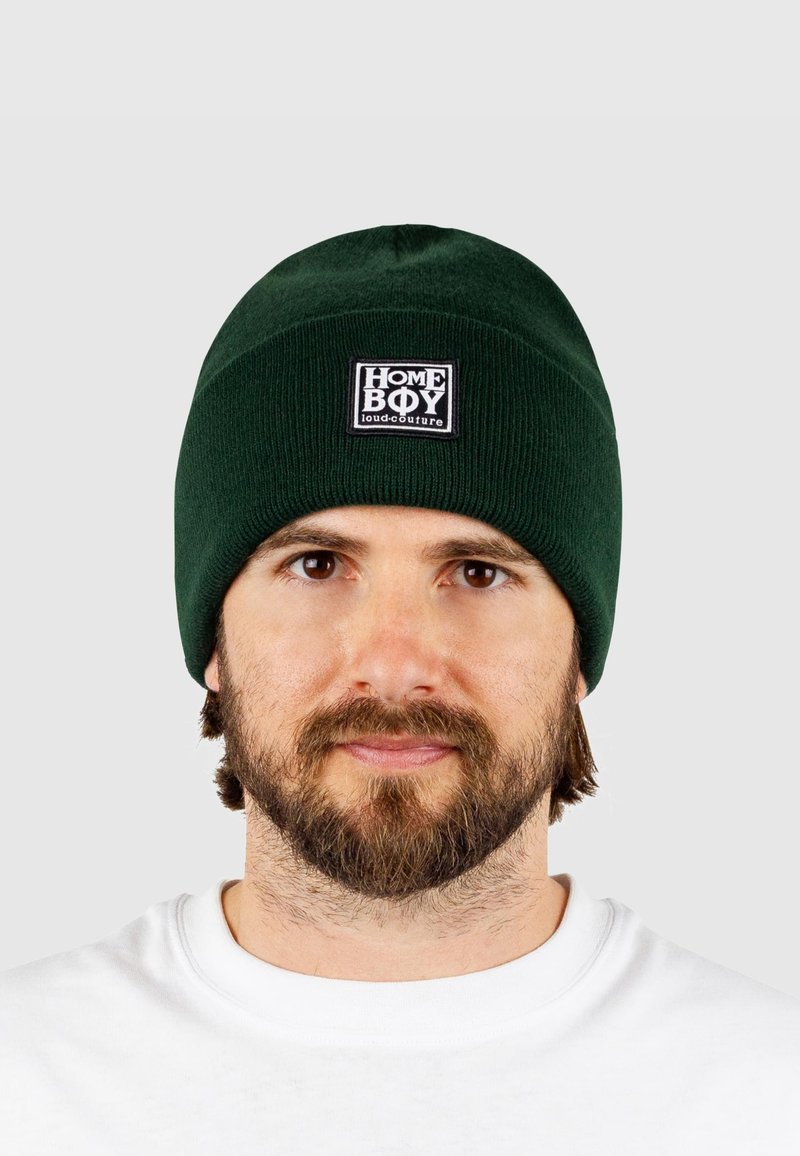 Berretto di lana verde lavorato a maglia con una texture a coste e un'etichetta nera quadrata con testo bianco. Indossato da una persona su uno sfondo neutro.