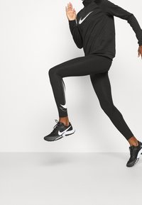 Mustat Nike-urheiluleggingsit, joissa on tiukka muoto ja valkoinen logo. Yhdistettynä samaan mustaan paitaan ja mustiin lenkkikenkiin, dynaamisessa asennossa.
