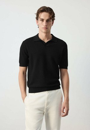 Joven con cabello ondulado que lleva una camiseta polo de punto negra de manga corta y pantalones de color crema claro, de pie frente a un fondo liso.
