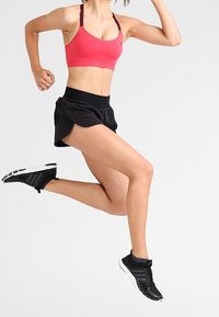 Vaaleanpunainen urheiluliivit, joissa säädettävät olkaimet, mustat kuvioidut shortsit ja mustat urheilukengät valkoisilla yksityiskohdilla, käytössä hypyn aikana.