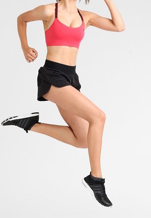 Rosa Sport-BH mit verstellbaren Trägern, schwarze, strukturierte Shorts und schwarze Sportschuhe mit weißen Akzenten, getragen während einer Sprungbewegung.