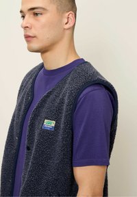 Námornícky modrá fleecová vesta s textúrovaným povrchom, s okrúhlym výstrihom a patentkami, cez fialové tričko s krátkymi rukávmi.