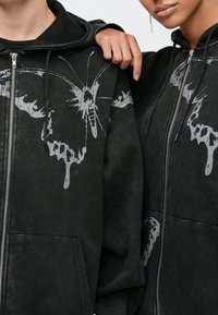 Μαύρα φούτερ με φερμουάρ και κουκούλα που διαθέτουν μεγάλα γκρι γραφικά πεταλούδας με σταγόνες. Μαλακή υφή, άνετη εφαρμογή και τσέπες καγκουρό.