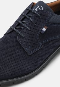 Zapato de ante azul marino con parte superior perforada, cordones planos, acentos cosidos y suela de goma texturizada. Incluye un sutil detalle del logo.