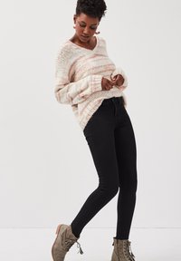 Suéter de gran tamaño, con cuello en forma de V en tejido de rayas crema y rosa, combinado con jeans negros ajustados y botas de tobillo verde oliva.