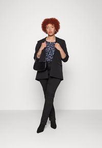Blazer negro sobre una blusa de tonos púrpura y negro con estampado, combinado con pantalones negros y un bolso negro texturizado; modelo posa con confianza.