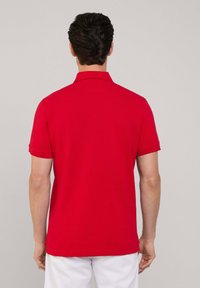 Polo rosso con colletto e maniche corte, caratterizzato da una texture liscia e un taglio dritto sul fondo. Nessun motivo o accentuazione visibile.