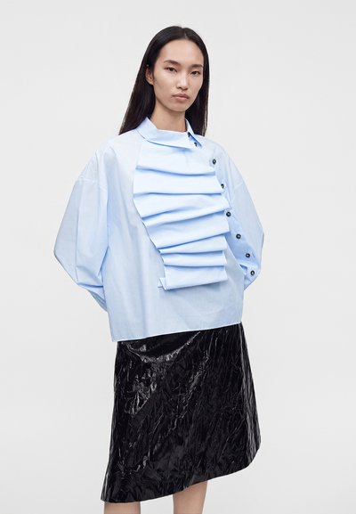 Femme portant un chemisier bleu clair oversize avec un détail plissé à l'avant et une jupe noire brillante texturée longueur genou sur un fond uni.