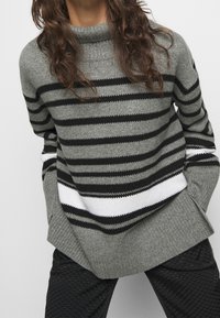 Szary sweter z dzianiny z czarno-białymi poziomymi paskami, kołnierzem w kształcie lejka i prążkowanymi mankietami. Połącz z fakturowanymi czarnymi spodniami.
