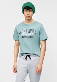 Νέος άνδρας που φοράει πράσινο σκουφάκι, ανοιχτό μπλε μπλουζάκι Jack & Jones και ανοιχτό γκρι φόρμα με μαύρο κορδόνι σε απλό φόντο.