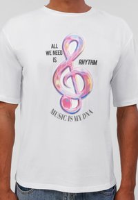 Balta kokvilnas T-kreklu ar krāsainu muzikālo piezīmi grafiku, ar melnu tekstu virs un zem tā, kas saka: "Mums viss, kas nepieciešams, ir ritms, Mūzika ir mans DNS."