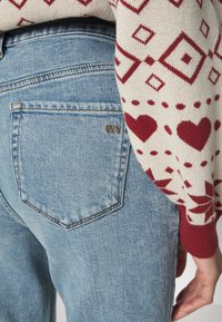 Os jeans azul claro apresentam um bolso traseiro padrão com um padrão de diamante costurado e o logótipo dourado "IVY". O suéter de malha tem um padrão geométrico vermelho.