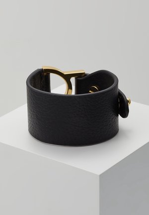 Bracelet manchette en cuir noir texturé avec une large bande et un fermoir en métal doré, présenté sur un socle blanc devant un fond gris.