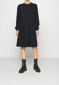 Kvinna som bär en svart långärmad knälång klänning i kombination med svarta kraftiga läder ankelskor mot en enfärgad bakgrund.