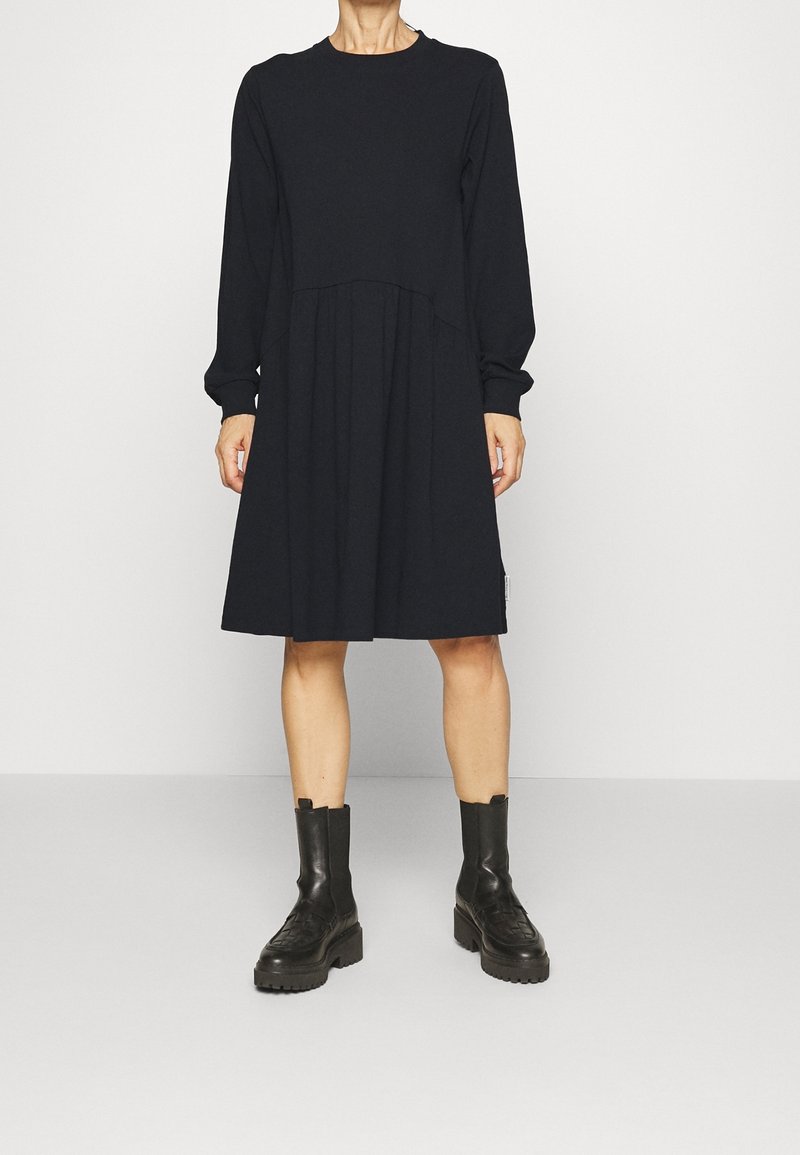 Kvinna som bär en svart långärmad knälång klänning i kombination med svarta kraftiga läder ankelskor mot en enfärgad bakgrund.