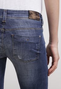 Jean skinny en denim bleu avec détails usés, étiquette en cuir marron à la taille, et poches arrière aux bords arrondis avec rivets métalliques.