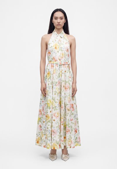 Femme portant une robe maxi sans manches, à col, ceinturée, avec un imprimé floral pastel et des chaussures en satin à bout pointu, debout devant un fond uni.