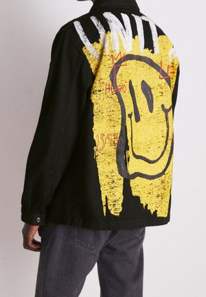 Persona con chaqueta negra que lleva un gran emoticono sonriente amarillo desgastado y texto blanco "UNITY" en la espalda, de pie frente a un fondo liso.