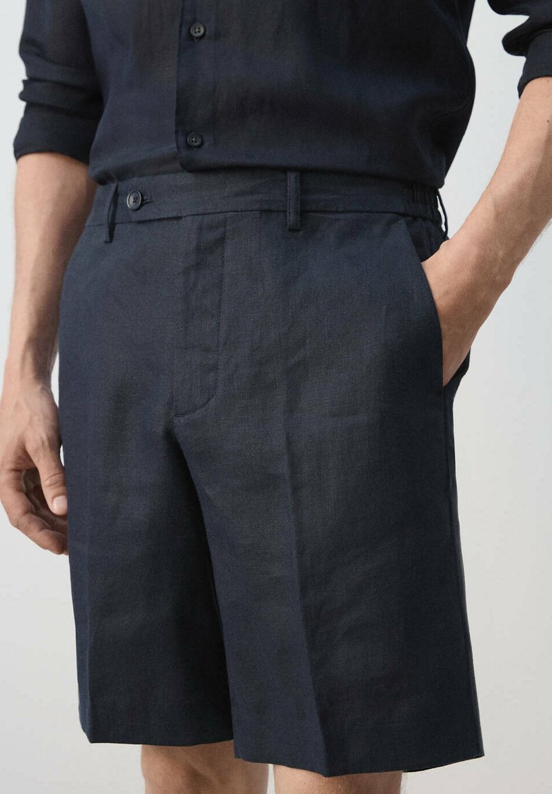 Homme portant un short en lin bleu marine foncé et une chemise boutonnée assortie, une main dans la poche, sur un fond uni.