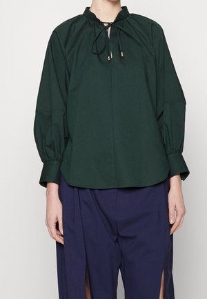 Vrouw draagt een donkergroene blouse met lange mouwen en striksluiting bij de hals en donkerblauwe wijde broek met voorsplitten.