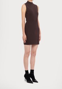 Brun ärmlös klänning med hög krage, figurnära passform, slät textur, kombinerad med svarta strumpor och spetsiga klackar.