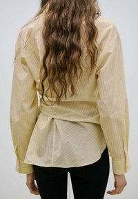 Oseba z dolgimi valovitimi rjavimi lasmi, oblečena v svetlo rumeno srajco z ozkimi črtami in črne hlače, prikazana od zadaj na enostavnem ozadju.