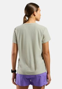 T-shirt de sport vert clair à manches courtes, texture lisse et logo dans le haut du dos. Porté avec des shorts violets.