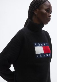 Musta poolokauluksinen neule, jossa on resorit hihansuissa. Edessä värikäs TOMMY JEANS -logopainatus punaisena, valkoisena ja sinisenä.