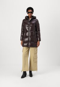 Bruine puffer jas met een glanzende afwerking, rits sluiting, capuchon, zijzakken en een logopatch, gedragen over beige cargobroeken en zwarte hoge hakken.