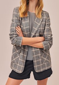 Femme portant un blazer à carreaux gris avec des manches retroussées, un haut plissé gris et un short noir avec ourlet retroussé, les bras croisés à la taille.