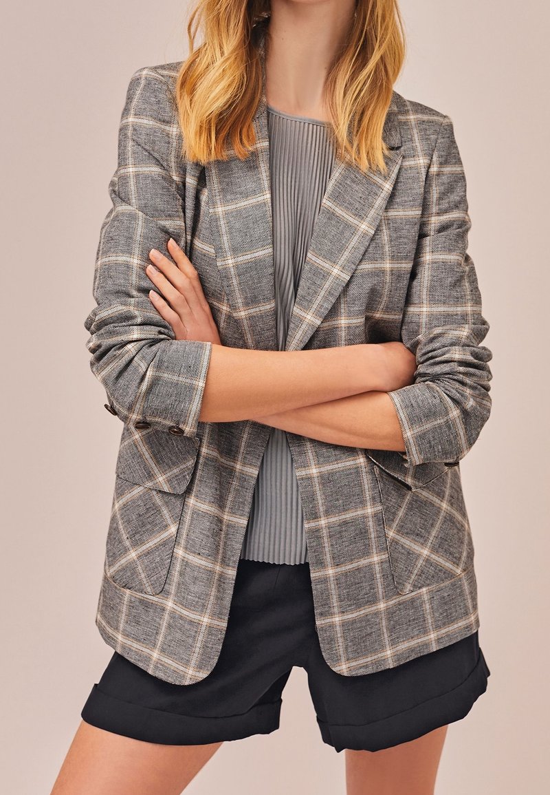 Femme portant un blazer à carreaux gris avec des manches retroussées, un haut plissé gris et un short noir avec ourlet retroussé, les bras croisés à la taille.