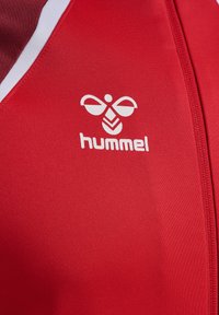 Rotes Sporttrikot mit weißen Akzenten. Besitzt ein auffälliges Hummel-Logo in Weiß auf der Brust. Hergestellt aus glattem, strukturiertem Stoff.