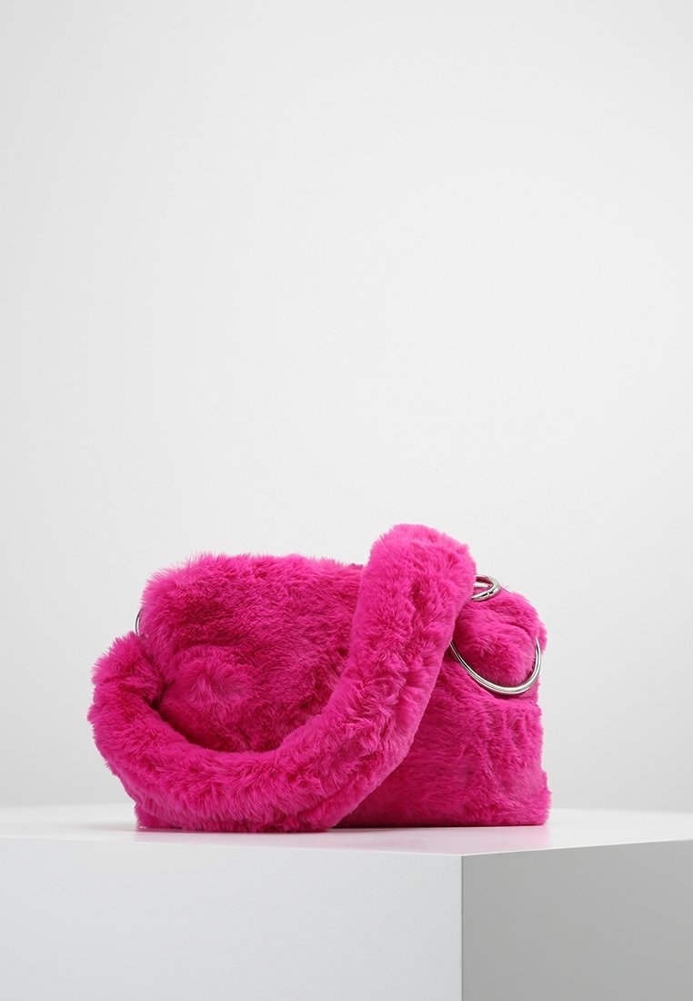 Felroze, pluizige handtas met een dikke, harige band en zilveren metalen ringdetail, tentoongesteld op een wit voetstuk tegen een neutrale achtergrond.