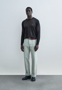 Άνδρας που στέκεται μπροστά σε απλό τοίχο, φοράει μαύρο μακρυμάνικο πουκάμισο, ανοιχτό γκρι παντελόνι με μανσέτες, κόκκινη ζώνη, μαύρα παπούτσια και κολιέ με χάντρες.