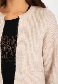 Cardigan tricoté rose clair avec une texture côtelée et une ouverture sur le devant. Il comporte une poche et est superposé sur un haut noir avec un motif de lion en sequins.
