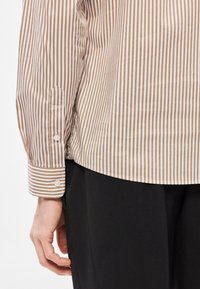 Prúžkovaná košeľa v béžovej a bielej farbe s hladkou textúrou, vybavená gombíkovými manžetami a zaobleným lemom, doplnená čiernymi nohavicami.