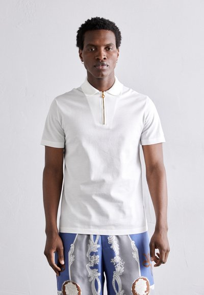 Hvid kortærmet polo-shirt med lynlås krave, der har en guld lynlås detalje. Stoffet ser glat og letvægts ud.