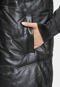 Czarny skórzany element garderoby o fakturowanym wykończeniu, z ręką wsuniętą w boczną kieszeń, prezentujący nowoczesny, elegancki design.