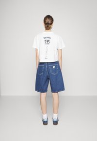 Hvit cropped T-skjorte med bakgrunnsgrafikk, paret med løse blå denimshorts. Shortsene har lommer og et logo-patch bak.
