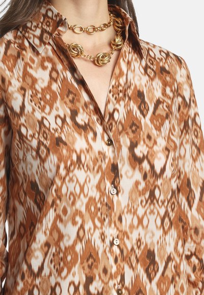 Blusa con motivo marrone e crema con colletto. Presenta bottoni sul davanti e una texture liscia. La collana a catena dorata esalta la scollatura.