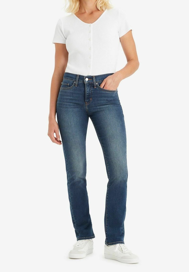 Femme portant un haut blanc à manches courtes boutonné, un jean slim bleu taille haute et des baskets blanches, debout avec une main dans la poche.
