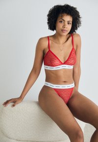 Set di lingerie rossa composto da un bralette trasparente e un perizoma abbinato, entrambi con un motivo testurizzato e una fascia elastica bianca con l'etichetta "GUESS."
