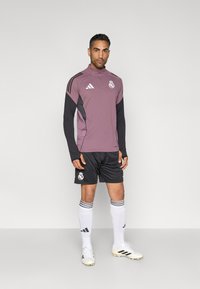 Μωβ και μαύρο μακρυμάνικο μπλουζάκι με φερμουάρ στον γιακά, μαύρο σορτς, λευκές κάλτσες με ρίγες και λευκά αθλητικά παπούτσια, με λογότυπο.