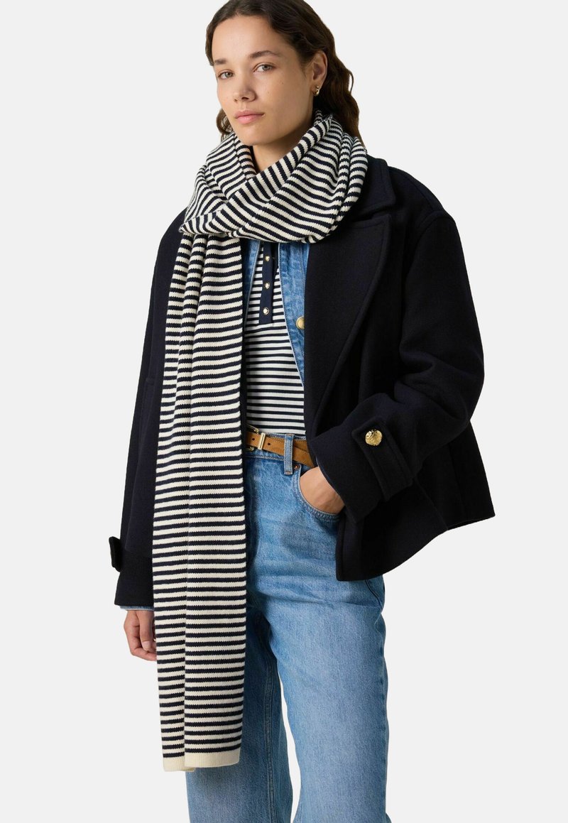 Écharpe rayée en noir et crème, faite de tissu doux, de forme longue et rectangulaire avec une texture tricotée épaisse et des extrémités de couleur crème.