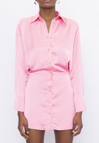 Rosa skjortklänning med lång ärm, tillverkad av ett mjukt material, med spetsig krage och fullknäppt fram. Kortskaft.