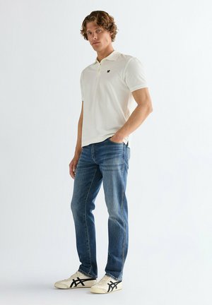 Junger Mann mit lockigem Haar, der ein weißes Polohemd, blaue Jeans und weiße Sneaker mit schwarzen Streifen trägt und mit einer Hand in der Tasche steht.