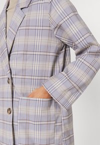 Main reposant dans la poche d'un blazer à carreaux beige et bleu clair avec des boutons marron, visible de la poitrine au poignet sur un fond neutre.