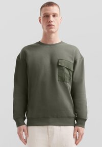 Sweter w kolorze oliwkowym, wykonany z mieszanki bawełny, z dużą naszywaną kieszenią na lewej piersi, okrągłym dekoltem oraz prążkowanymi mankietami i dołem.