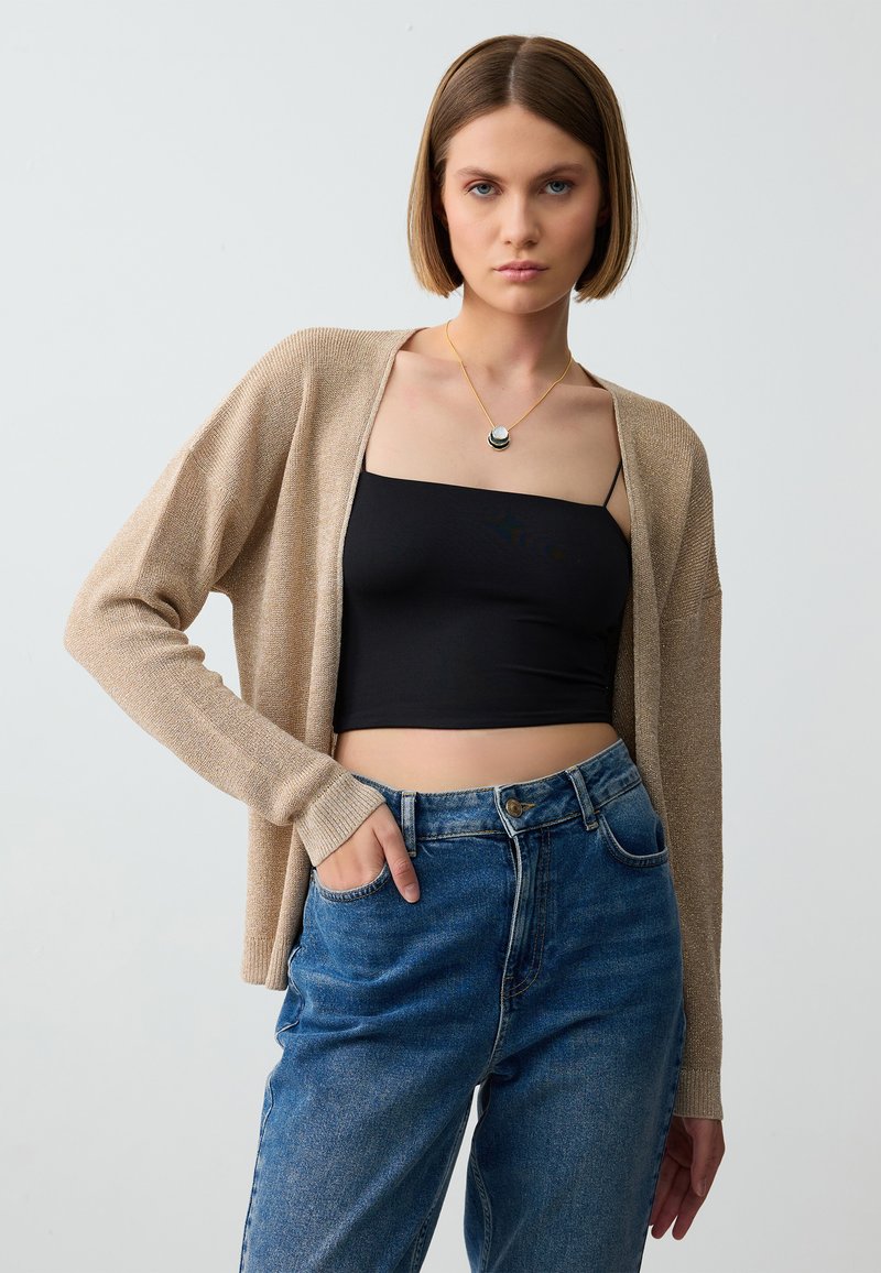 Beige gestrickte Strickjacke mit langen Ärmeln, getragen über einem schwarzen taillierten Oberteil. Das Model posiert mit den Händen in den Taschen einer blauen hoch geschnittenen Jeans.