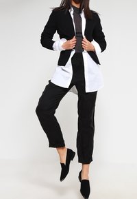 Blazer noir ajusté sur une chemise blanche à boutons, associé à un pantalon noir ample et des ballerines noires. Le tissu texturé crée un contraste entre les couches.